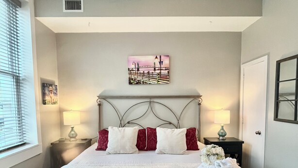 A neatly made bed with white bedding and red pillows, a headboard with a decorative design, a wall-mounted artwork, and a bedside table with a lamp. - Perfect Work Retreat on Market St. Downtown ILM (Wilmington)