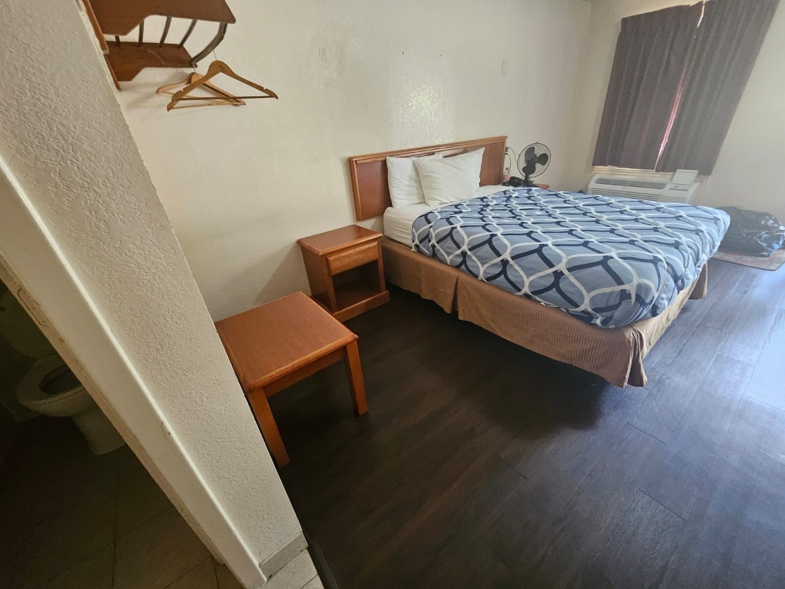A bedroom with a bed, a bedside table, a chair, a hanging coat rack, and a window with curtains.