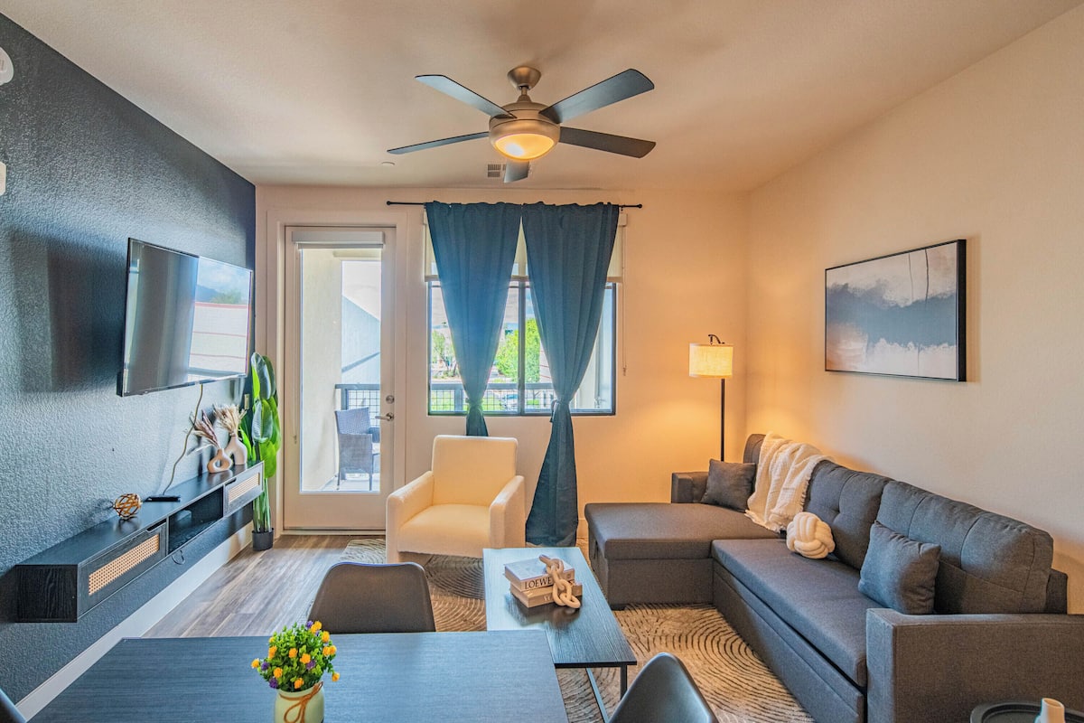 A modern living room with a grey sofa, a white armchair, a dining table with a vase of flowers, and a ceiling fan.