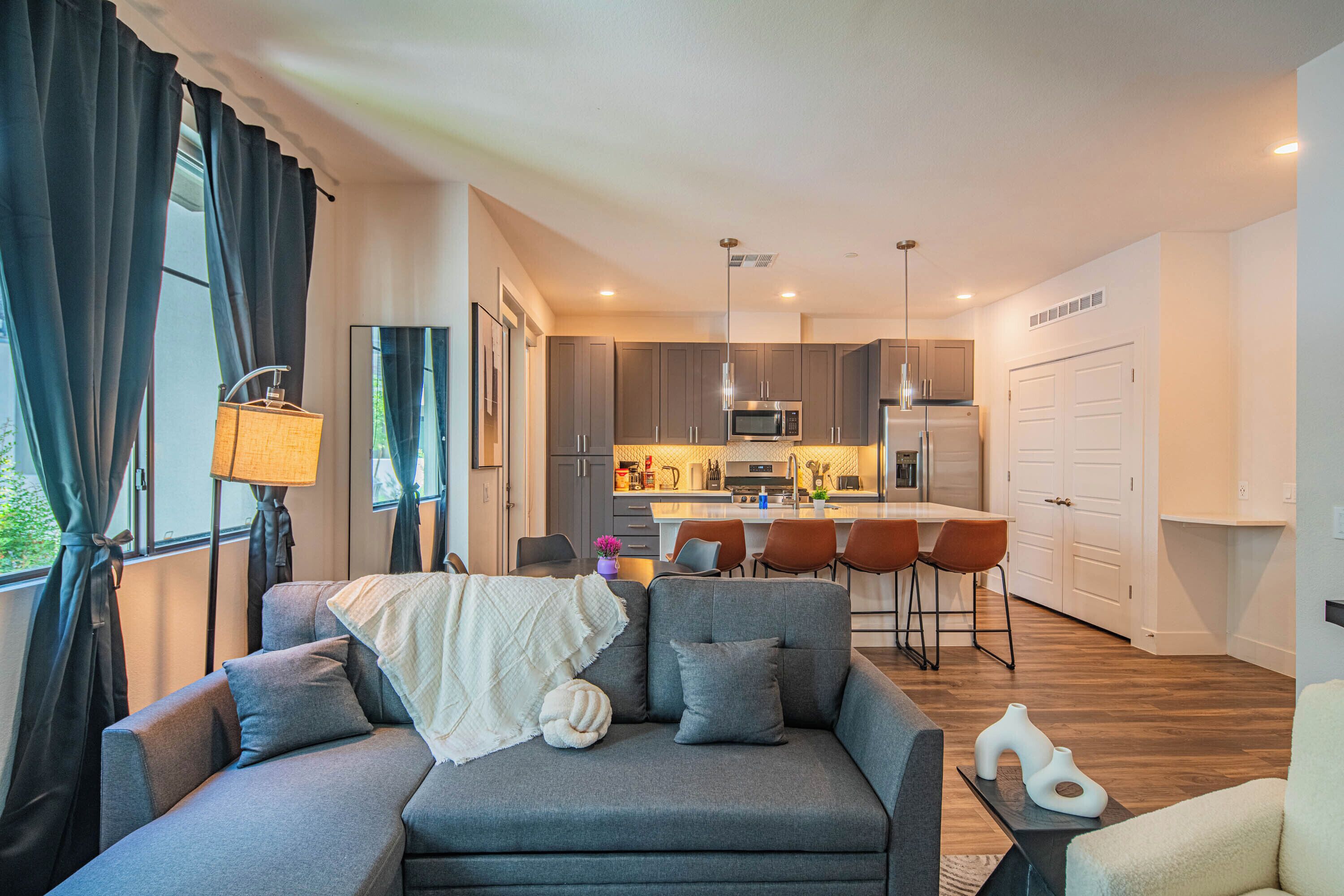 Una sala moderna con un sofá gris, una mesita y una lámpara. Se ve una cocina con microondas y refrigerador al fondo.