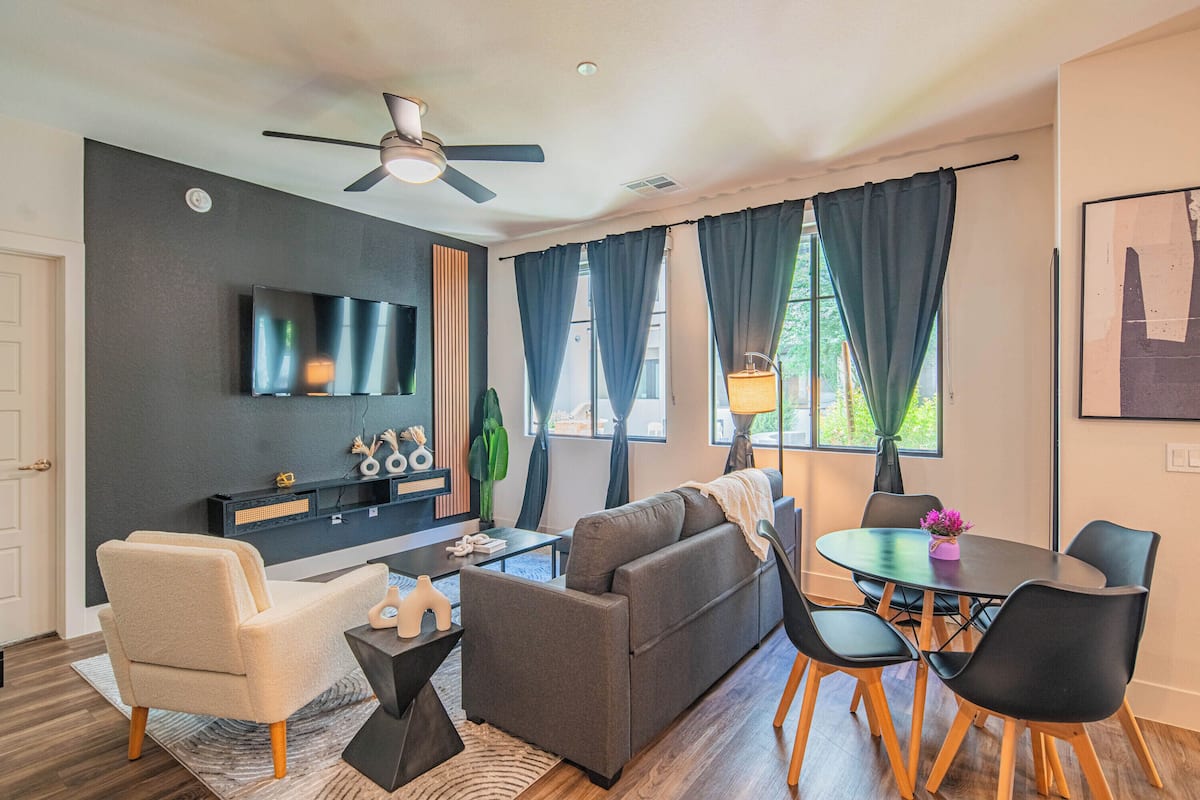 A modern living room with a grey sofa, a white armchair, a dining table with chairs, a ceiling fan, and a TV mounted on the wall.