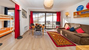 A cozy living room with a wooden bunk bed, a dining table with chairs, a sofa with cushions, a patterned rug, and a large window with a view of a snowy landscape. - Baikonour by Interhome (Le Corbier)