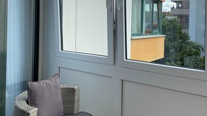 Een balkon met een rieten stoel en kussens, grote ramen en uitzicht op gebouwen en bomen.