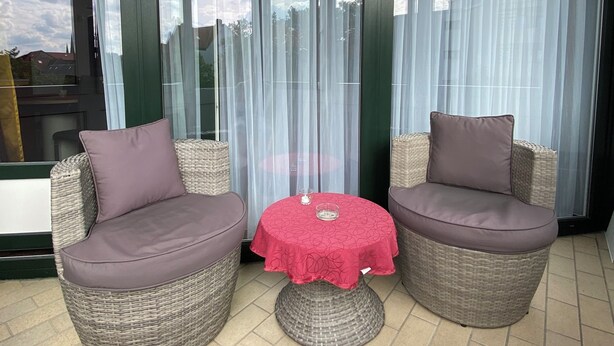 Een balkon met rieten meubilair, een klein tafeltje en dunne gordijnen.