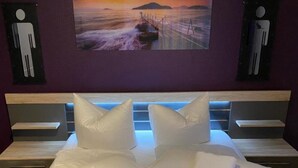 Een bed met witte lakens en kussens, een schilderij van een zonsondergang boven water, en twee aan de muur bevestigde pictogrammen voor een toilet en een douche.