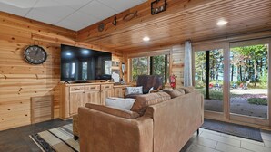 Ein gemütliches Blockhaushaus-Interieur mit Holzvertäfelung, einem großen Flachbildfernseher, einer Couch und Blick ins Freie.