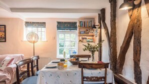 A cozy dining area with a table set for two, surrounded by wooden chairs. A bookshelf filled with books is visible in the background, and a window allows natural light to enter the room. - Blythburgh Cottage, Yoxford (Yoxford)