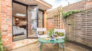 A cozy outdoor seating area with a table and chairs, a glass door leading to a room with a bed, and a brick wall with a wooden trellis. - Blythburgh Cottage, Yoxford (Yoxford)