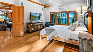Una camera da letto con un letto, una cassettiera in legno, una televisione e una finestra vista alberi e montagne.