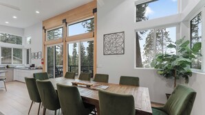 A modern dining area with a wooden table and green upholstered chairs, large windows, and a view of trees. - Yosemite Park / Forest View, Patio & BBQ Area (TUOLUMNE MEADOWS)