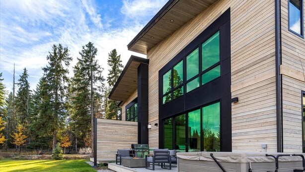A modern house with large windows, a patio with outdoor furniture, and a forested backdrop. - Whitefish Vista Retreat (Whitefish)