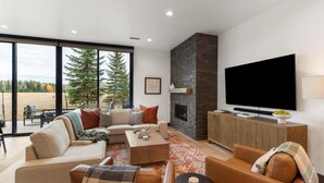 A modern living room with a fireplace, a large TV, and a view of trees and a field. - Whitefish Vista Retreat (Whitefish)