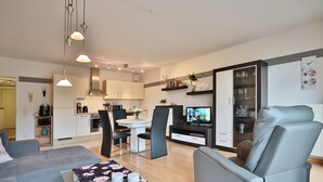 A modern living room with a sofa, a dining table with chairs, a TV, and a kitchen area with cabinets and appliances.