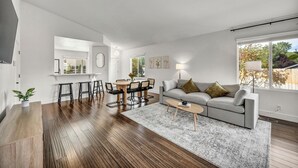 Ein modernes Wohnzimmer mit grauem Sofa, Holzboden, einem Essbereich mit Tisch und Stühlen sowie einem großen Fenster für natürliches Licht.