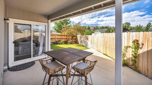 Eine überdachte Terrasse mit Holztisch und Korbstühlen, eine Schiebetür und Blick auf einen Garten mit Holzzaun und Grünflächen.