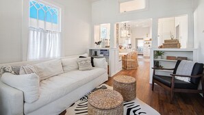 A spacious living room with a white sofa, wicker ottomans, a zebra-patterned rug, and a dining area with a wicker chair and table. - Historic Cottage in Olde Town BSL - One Block from Beach & Dining (Bay St. Louis)