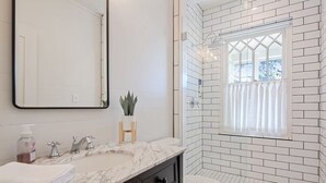 A bathroom with a white tiled shower, a marble sink, and a toilet. - Historic Cottage in Olde Town BSL - One Block from Beach & Dining (Bay St. Louis)