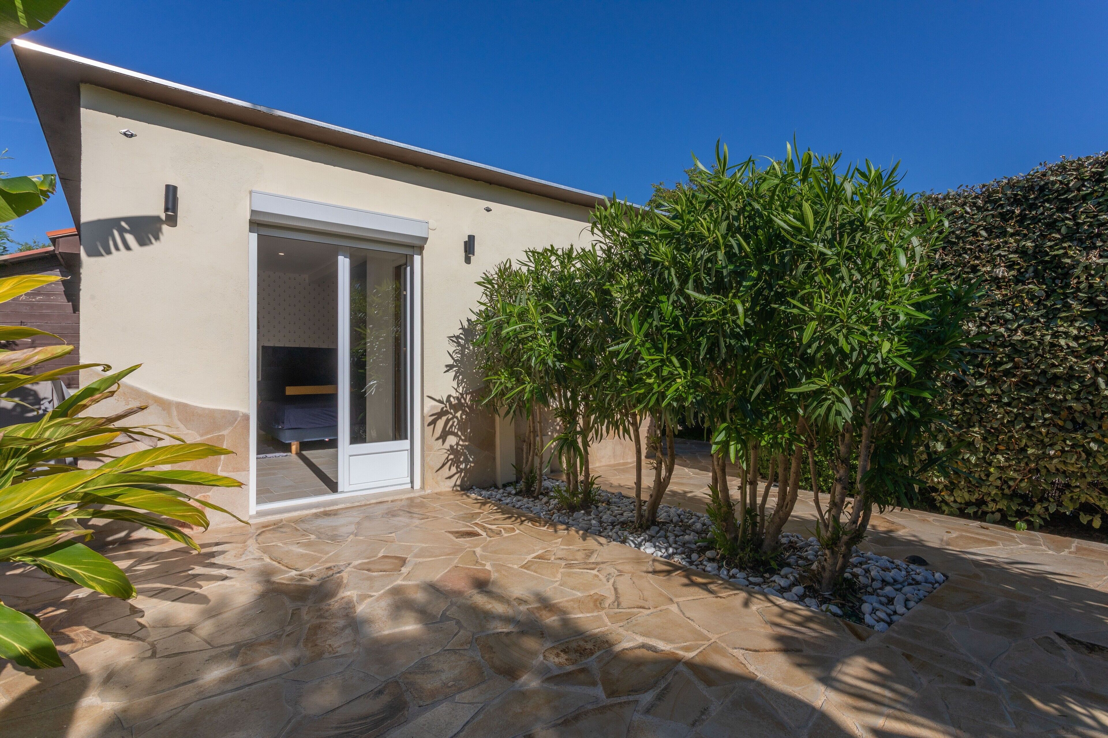 Un bâtiment moderne doté d’une terrasse en pierre, d’une entrée avec une porte en verre et d’un petit jardin agrémenté de plantes.