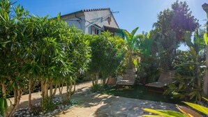 Ein steiniger Pfad führt zu einem Haus mit einem Ziegeldach, umgeben von üppiger Vegetation und einer kleinen Sitzecke mit einem Stuhl und einem Tisch.