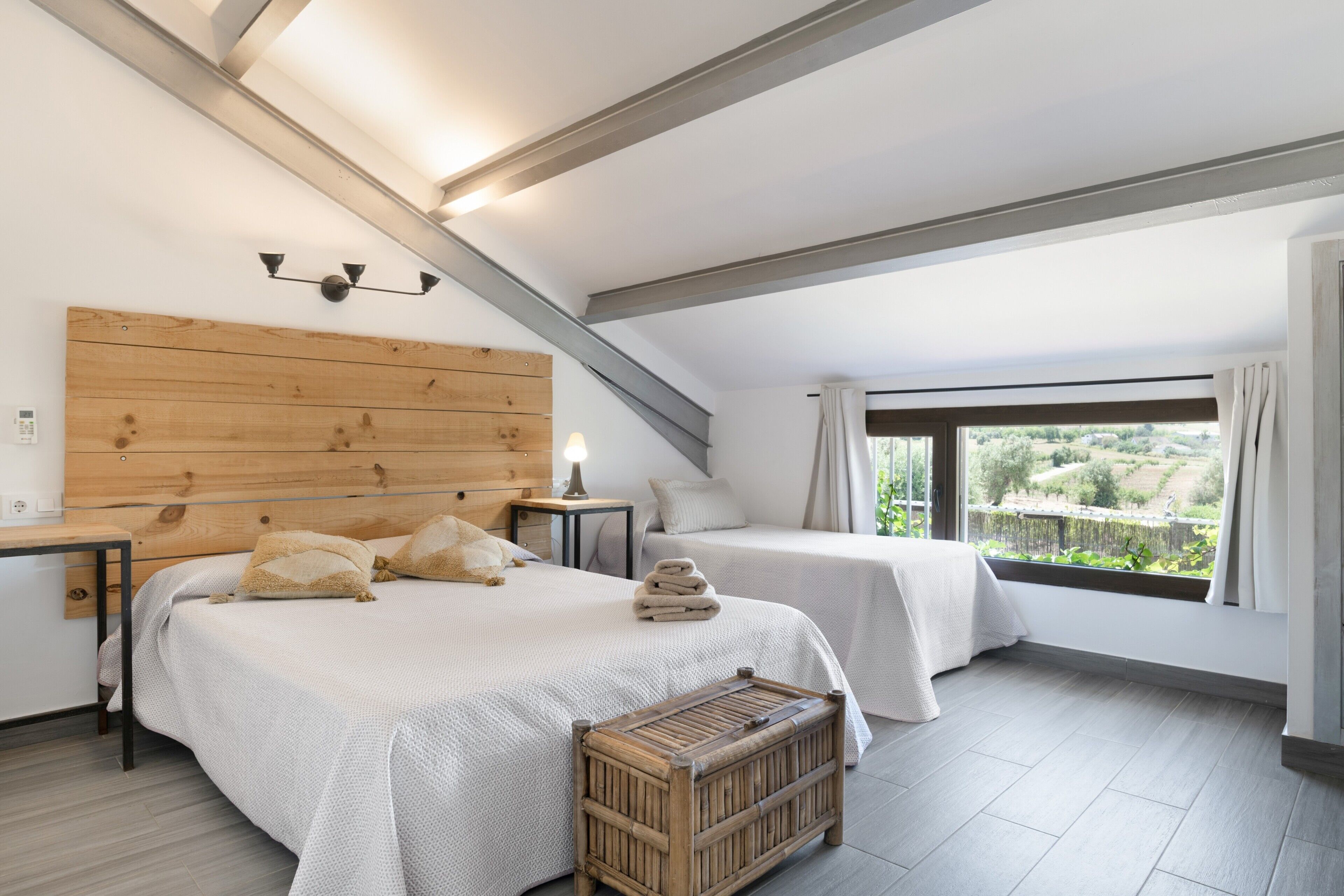 A bedroom with a sloped ceiling, two beds, a wooden headboard, a nightstand, a bedside lamp, and a view of greenery outside.
