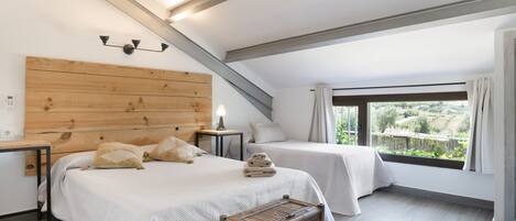 A bedroom with a sloped ceiling, two beds, a wooden headboard, a nightstand, a bedside lamp, and a view of greenery outside.