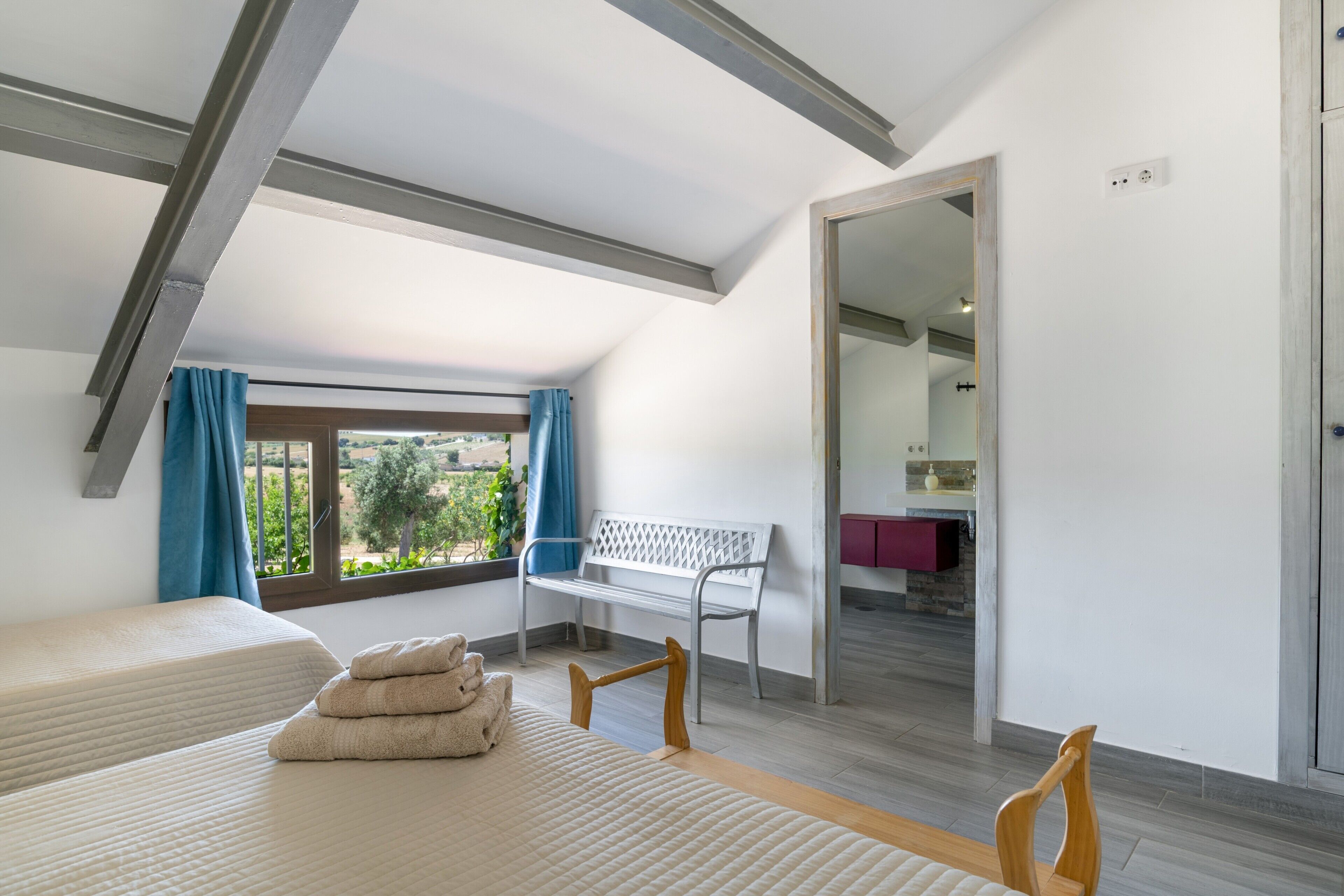 Une chambre avec un plafond de style industriel, une fenêtre avec des rideaux, un banc, un lit avec un oreiller et une salle de bain avec un miroir et un lavabo.