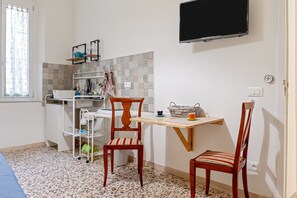 A small room with a tiled floor, a desk with a chair, a television mounted on the wall, and a window with curtains.