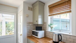 Una cocina con armarios blancos, microondas, hervidor y ventana con persianas.