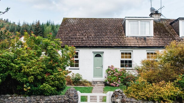 Una casita blanca con puerta verde, paredes de piedra y una verja de piedra.