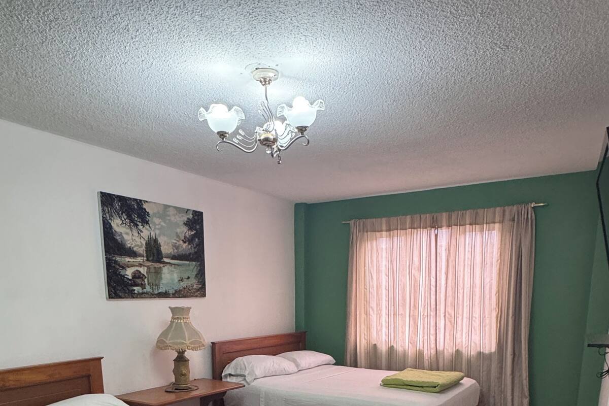 A room with two wooden beds, a nightstand with a lamp, a painting on the wall, and a window with curtains.