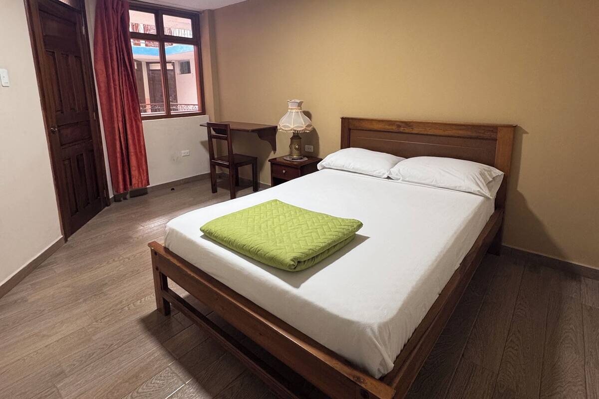 A neatly made bed with a green blanket, a wooden headboard, a nightstand with a lamp, and a window with curtains.