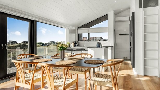 Ein moderner Essbereich mit großem Tisch und Stühlen, eine Küche im Hintergrund und ein Blick auf die Landschaft durch große Fenster.