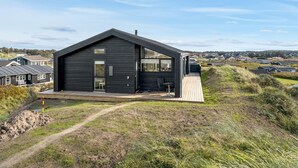 Ein modernes schwarzes Haus mit einer Terrasse, umgeben von einer grünen Landschaft und Wohnhäusern im Hintergrund.