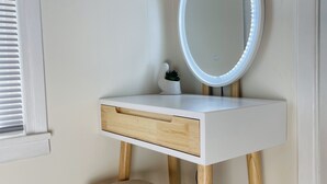 En vit sminkbord med ett runt spegel och LED-lampor, en liten krukväxt och en träpall.