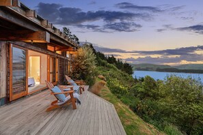 A wooden cabin with a deck, outdoor seating, and a view of a lake and hills.