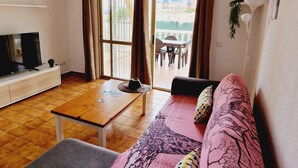 Ein Wohnzimmer mit Fernseher, Tisch, Sofa und einem Balkon mit Tisch und Stühlen.