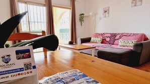 Ein Hotelzimmer mit einem Holztisch, einer Couch und einem Fenster mit Vorhängen. Auf dem Tisch liegen Prospekte und am Fensterbrett steht eine Pflanze.