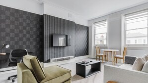 A modern living room with a sofa, armchair, and a coffee table. A TV is mounted on the wall, and there is a window with a view of buildings. - Cool Notting Hill 1BR, Fully Furnished & Ready, by Blueground (London)