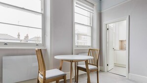 A room with two wooden chairs and a round table, large windows with blinds, a radiator, and a view of buildings outside. - Cool Notting Hill 1BR, Fully Furnished & Ready, by Blueground (London)
