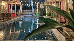 A pool area with a clear view of the water, surrounded by wooden structures and palm trees, under string lights. - The Bangka Place (Enrique Villanueva)