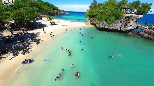 A beach with clear turquoise water, people swimming and sunbathing, and a rocky outcrop with a hut. - The Bangka Place (Enrique Villanueva)