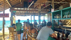 A beachside bar with patrons seated at tables, a bar area with bottles, and a sign indicating a cocktail zone. - The Bangka Place (Enrique Villanueva)