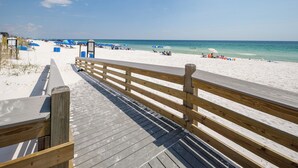 Ein Holzsteg führt zu einem Strand mit Sonnenschirmen und Liegestühlen.
