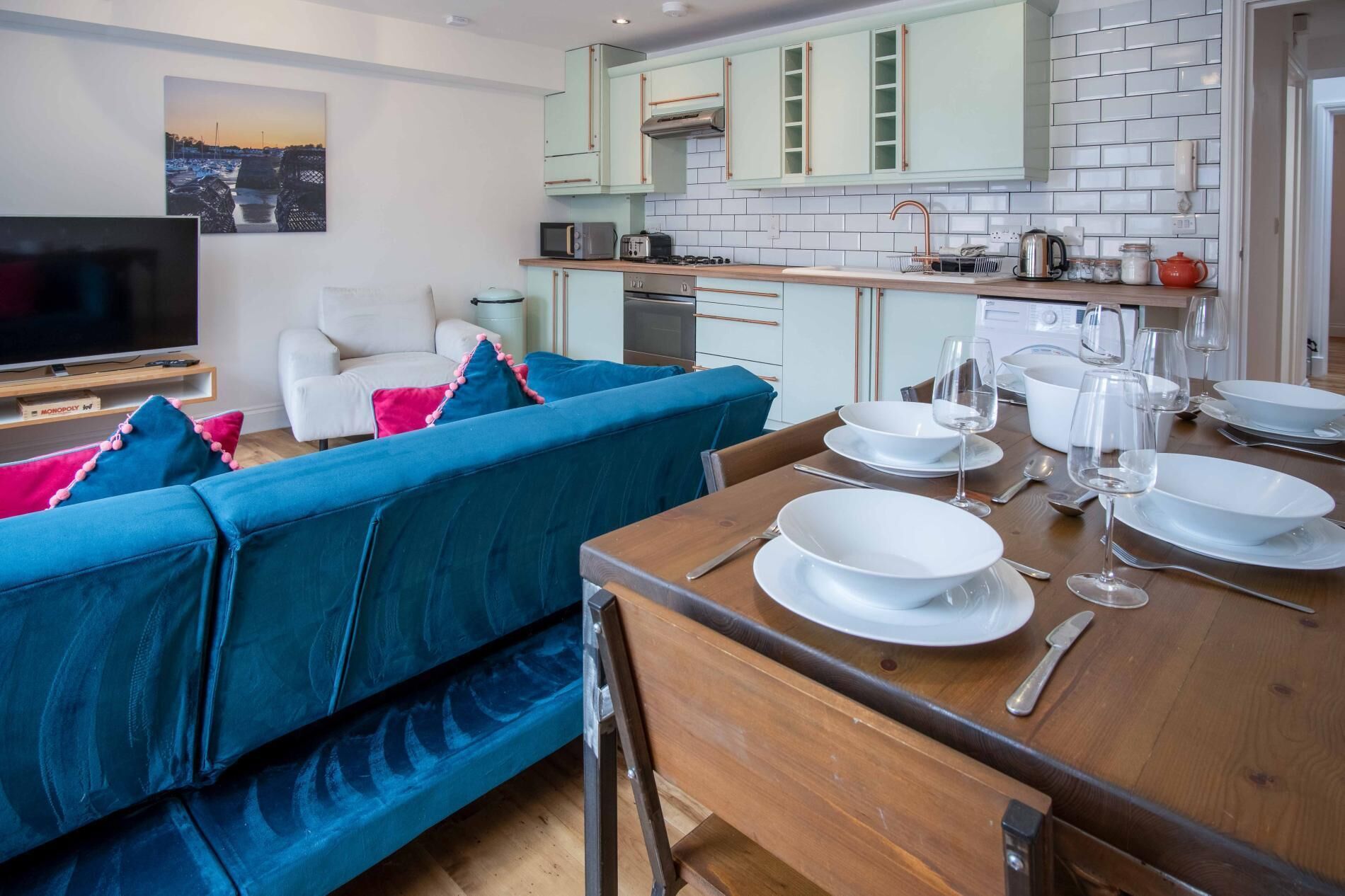 Ein modernes Wohnzimmer mit einem blauen Sofa, einem Holzespeisesaal mit weißen Tellern und Gläsern, und einer Küche im Hintergrund mit hellgrünen Schränken.