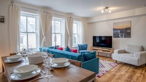Una sala moderna con una mesa de comedor de madera lista para una comida, un sofá azul, un sillón blanco y un televisor de pantalla plana.