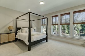 Ein Himmelbett mit dunklem Gestell, ein Nachttisch mit Lampe und Blick auf Bäume durch große Fenster mit Bambusrollos.