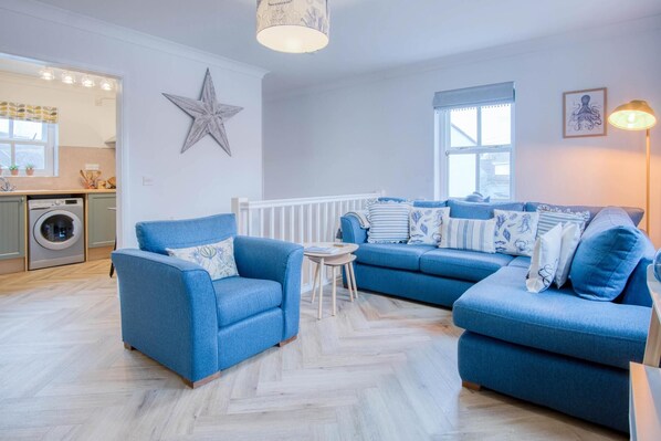 A living room with a blue sofa set, a star decoration on the wall, and a washing machine in the background. - Kilmore House - 2 Bedroom Apartment - Tenby (Tenby)