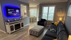 A modern living room with a flat-screen TV, a sofa, a coffee table, and a patterned ottoman. - Modern & Clean, Family Friendly, 1st Floor Condo (Rehoboth Beach)