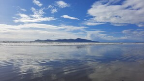 A beach with calm waters reflecting the sky, distant mountains, and a clear horizon. - Sunset cottage Paekākāriki - sand, sea and sunsets (Paekakariki)