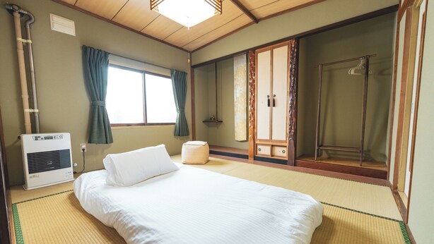 Ein traditioneller japanischer Raum mit Tatami-Boden, einem Bett mit weißer Bettwäsche, einem Heizgerät, einem Fenster mit Vorhängen und einer Schiebetür zu einem Duschbereich.